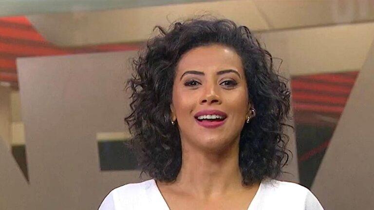 Apresentadora sorri durante programa de TV em estúdio com cenário em tons de vermelho e bege
