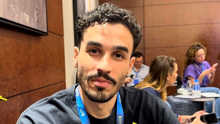 Homem com crachá azul posa durante evento em sala de conferências com pessoas ao fundo