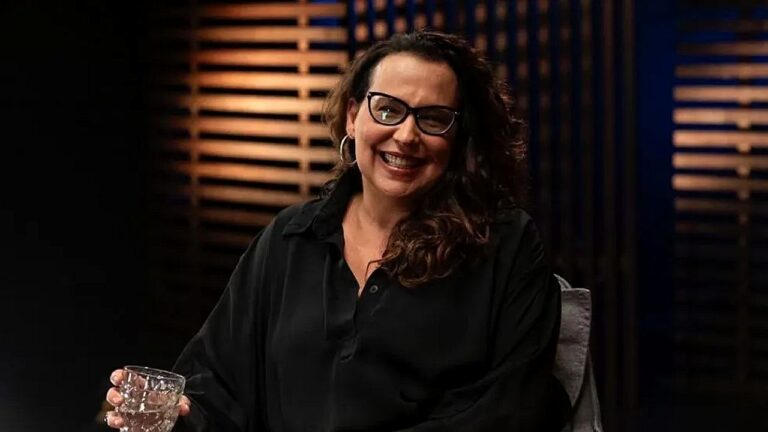 Mulher de óculos sorri durante entrevista em programa de TV com iluminação baixa no estúdio