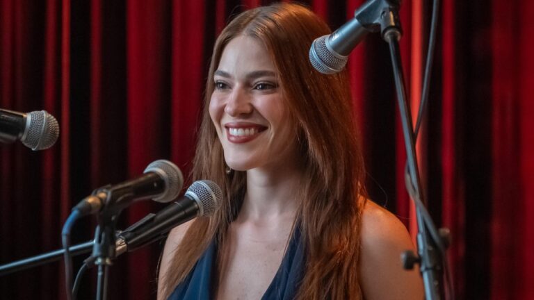 Ana Clara sorrindo diante de microfones em palco com cortinas vermelhas durante chamada do Estrelas da Casa