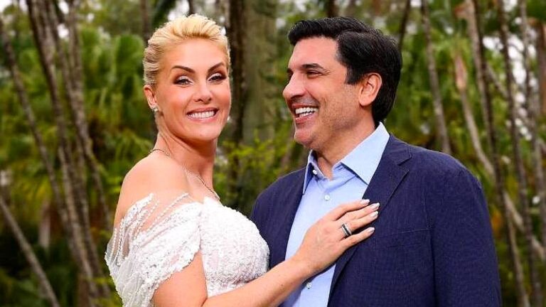 Casal sorri durante ensaio ao ar livre com vestido de noiva e terno azul em cenário com árvores