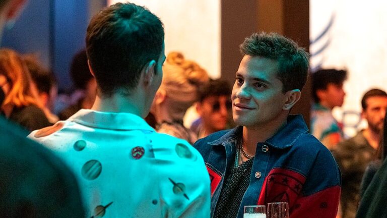 Dois homens conversam durante festa ou evento noturno com iluminação colorida ao fundo