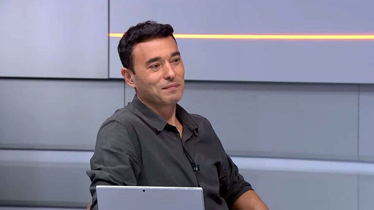 Homem de camisa cinza sentado em estúdio de TV ao lado de notebook com expressão sorridente