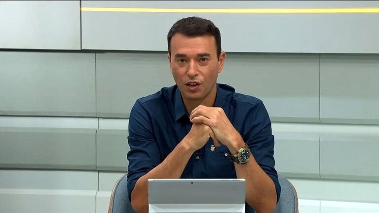 Homem de camisa azul apresenta programa jornalístico sentado diante de tablet em estúdio de televisão