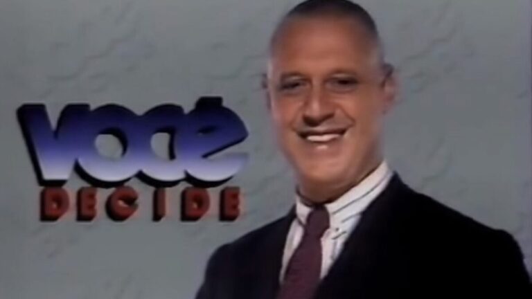 Antonio Fagundes de terno e gravata sorri diante do logotipo do programa Você Decide em fundo claro