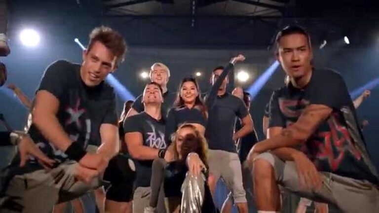 Grupo de jovens posa em palco iluminado durante apresentação de dança com refletores ao fundo