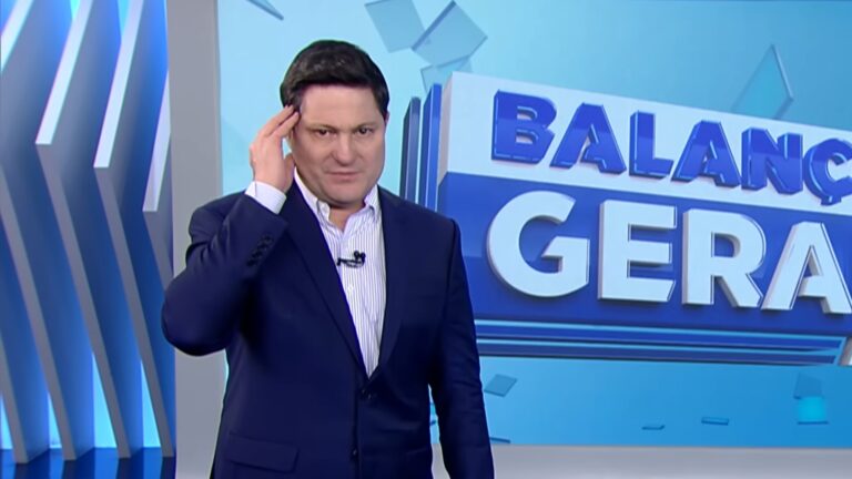 Eleandro Passaia, apresentador do Balanço Geral, veste um terno azul e apresenta o jornalístico em um cenário moderno, com barras laterais e um telão com o logotipo do programa da Record.