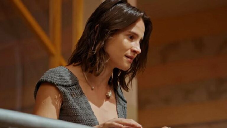 Mulher de cabelo curto inclina-se sobre mesa com expressão séria em cena de novela ambientada em interior de casa