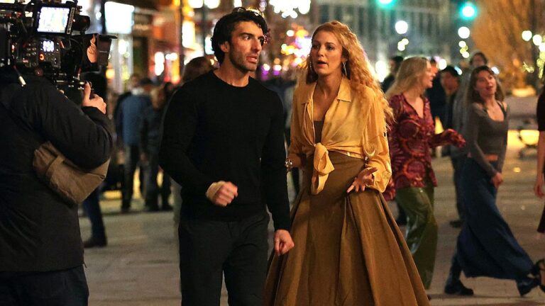 Homem e mulher caminham e conversam à noite em rua movimentada enquanto equipe grava cena com câmera profissional