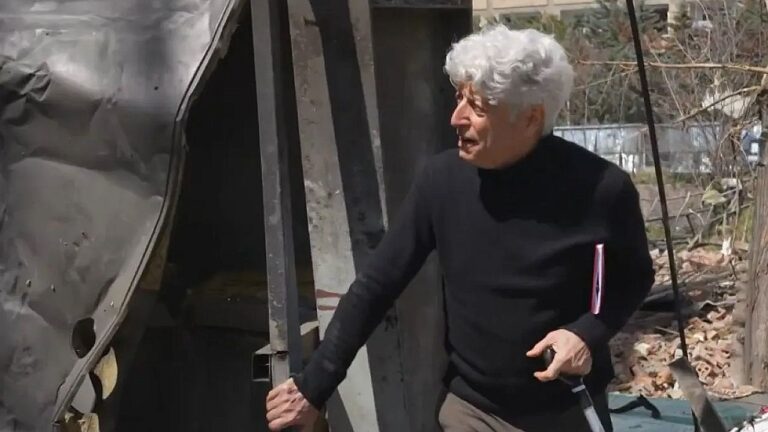 Homem idoso de cabelo branco abre porta de estrutura danificada em área com destroços ao redor