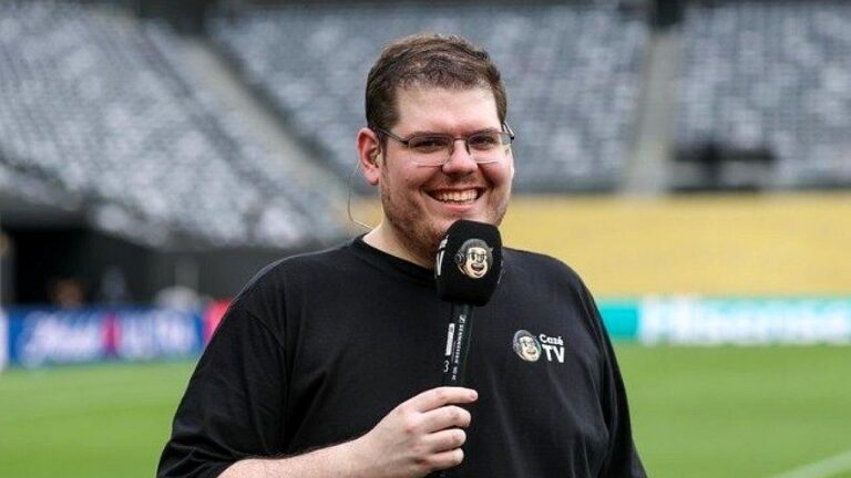 Repórter esportivo sorri no gramado de estádio segurando microfone da Cazé TV