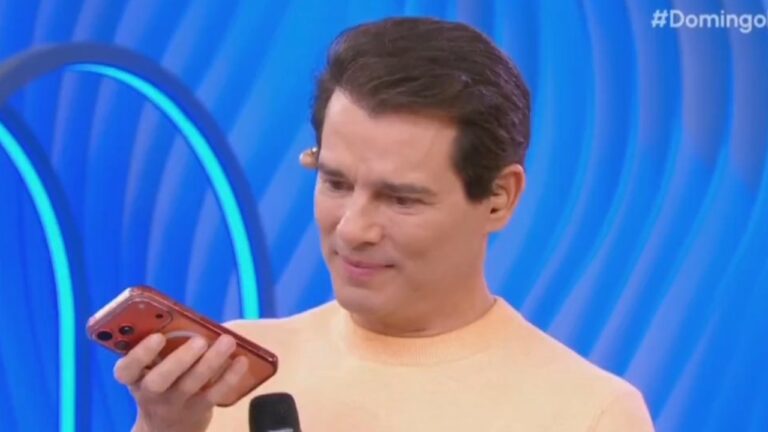 Celso Portiolli segura celular vermelho e observa a tela com expressão concentrada no palco do Domingo Legal no SBT