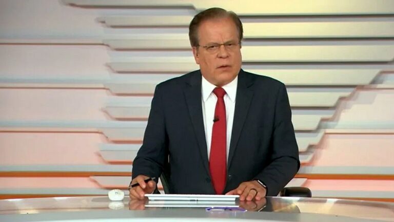 Homem de terno e gravata vermelha apresenta telejornal em bancada de estúdio de notícias