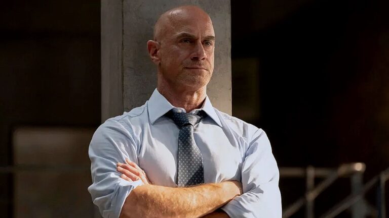 Texto Alternativo disse: Homem careca de camisa social e gravata cruza os braços com expressão séria em cena de série policial