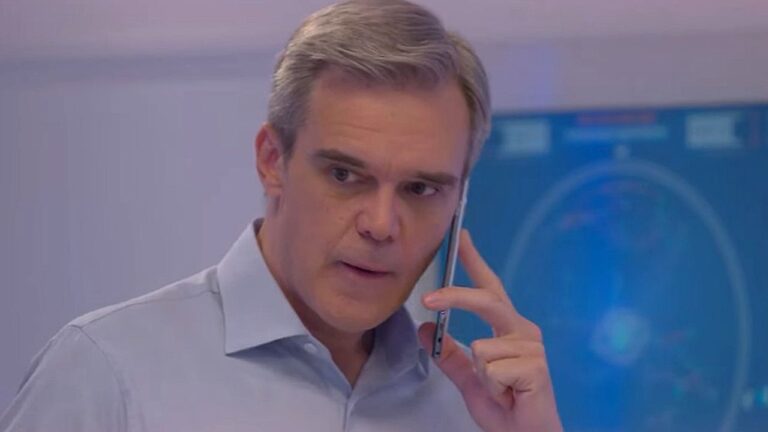 Homem de cabelo grisalho fala ao celular com expressão séria em cena de novela brasileira