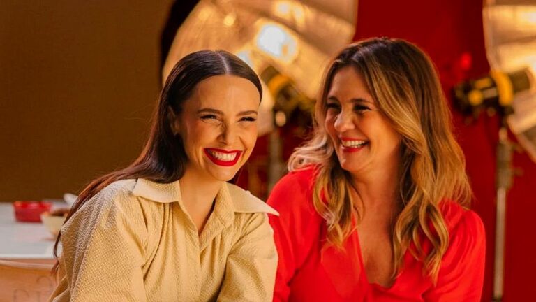 Duas mulheres sorrindo e conversando em estúdio com iluminação quente e equipamentos de gravação ao fundo