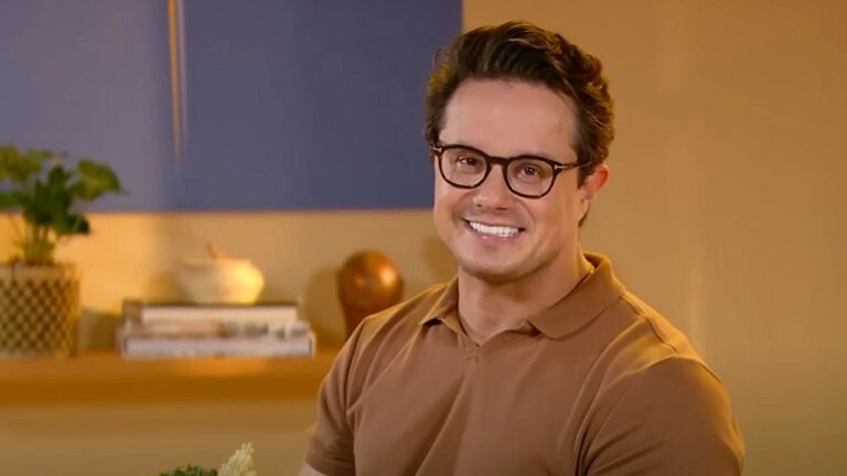 Homem de óculos e camisa marrom sorri sentado em ambiente interno com decoração ao fundo