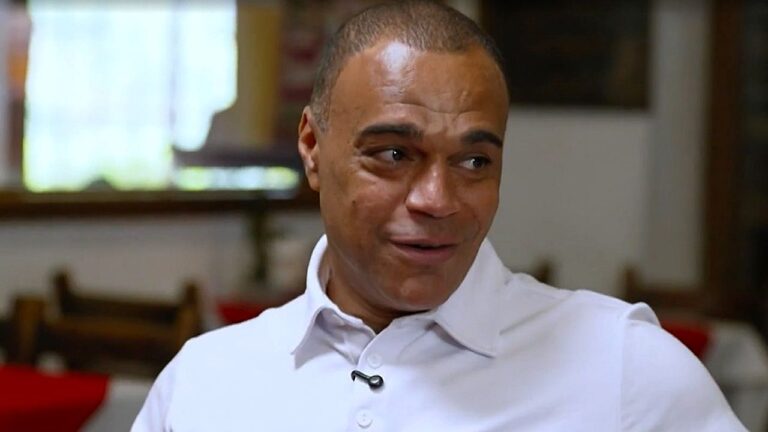 Denílson durante entrevista em restaurante usando camisa branca e microfone preso à gola