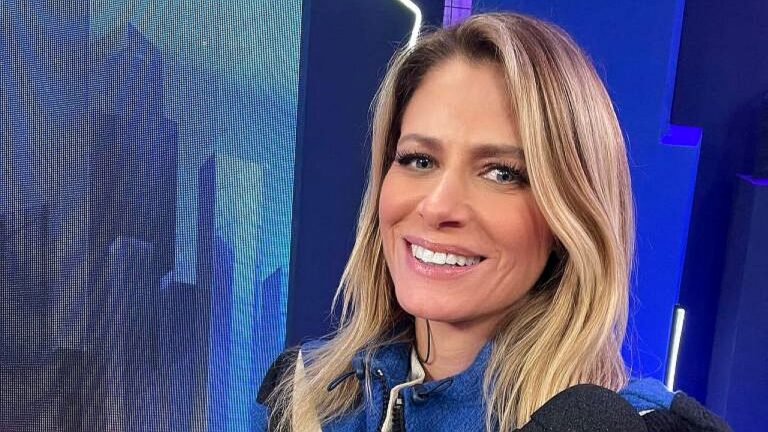Apresentadora loira sorri diante de câmera em estúdio com iluminação azul ao fundo