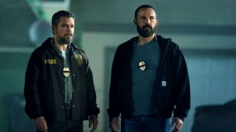 Dois homens com distintivos investigativos observam cena em ambiente escuro de thriller policial