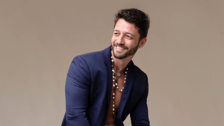 Diogo Venturieri sorri usando blazer azul aberto e colares posando em ensaio com fundo neutro