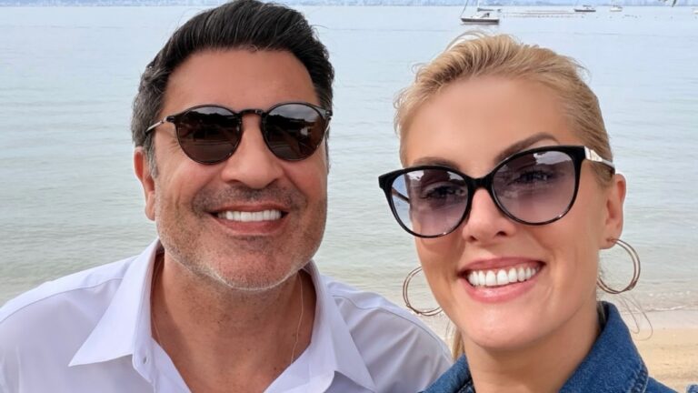 Edu Guedes e Ana Hickmann sorriem usando óculos escuros em selfie na praia com mar calmo e barcos ao fundo
