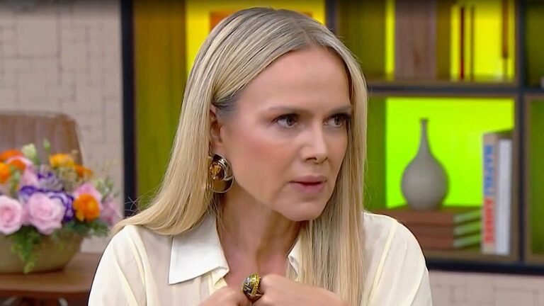 Mulher loira com brincos dourados fala durante entrevista em programa de TV com estante ao fundo