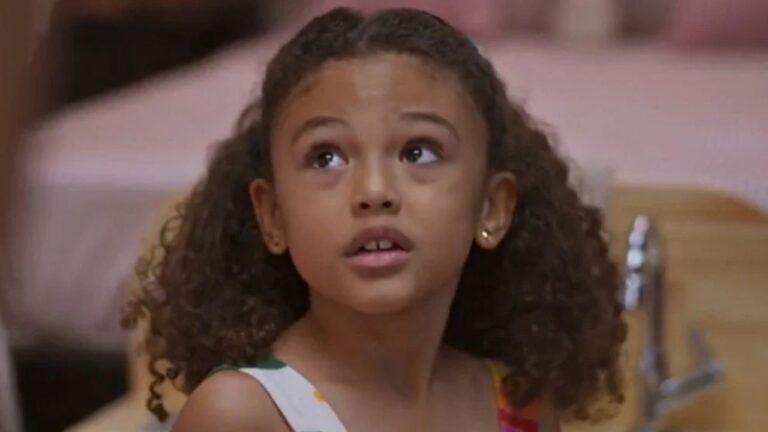 Menina com cabelos cacheados olha para cima com expressão curiosa em cena interna de novela