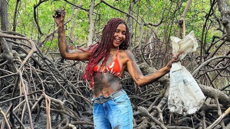 Emelly Souza sorrindo em área de manguezal com corpo coberto de lama e saco plástico nas mãos durante ação ambiental