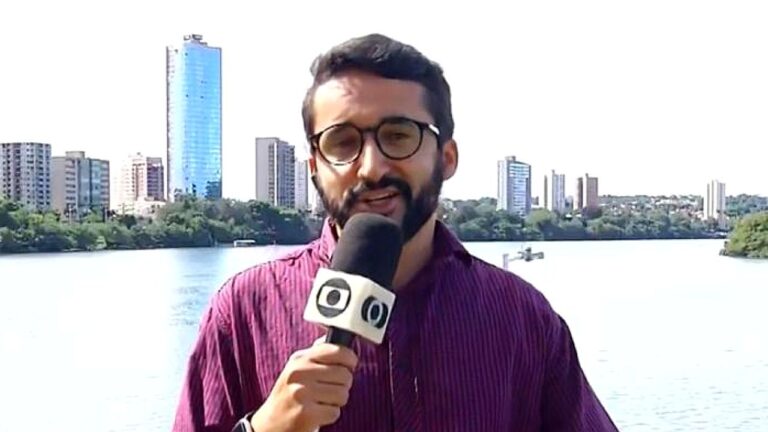 Repórter Emerson Ferreira segura microfone da Globo durante entrada ao vivo com lago e prédios ao fundo