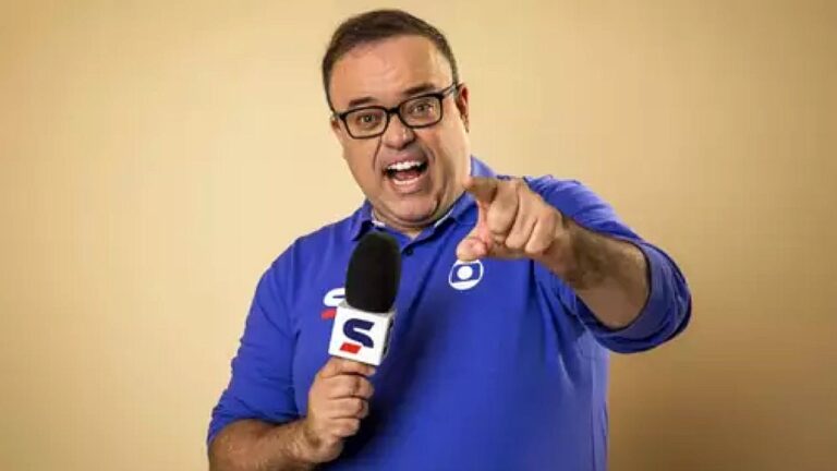 Homem de camisa azul da Globo segura microfone esportivo e aponta para a câmera sorrindo