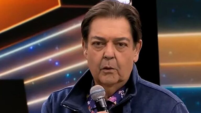Fausto Silva segura microfone durante participação em programa de televisão com cenário iluminado ao fundo