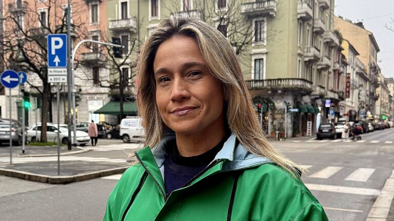 Mulher de casaco verde posa em rua urbana com prédios e carros ao fundo