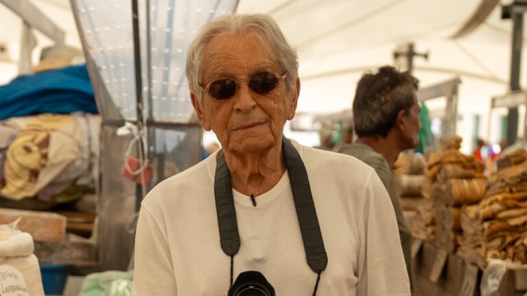 Fernando Gabeira usando óculos escuros e câmera pendurada no pescoço durante visita a mercado popular ao ar livre