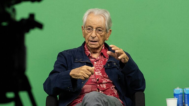 Fernando Gabeira fala gesticulando com as mãos sentado em cadeira durante evento com fundo verde e garrafa de água ao lado
