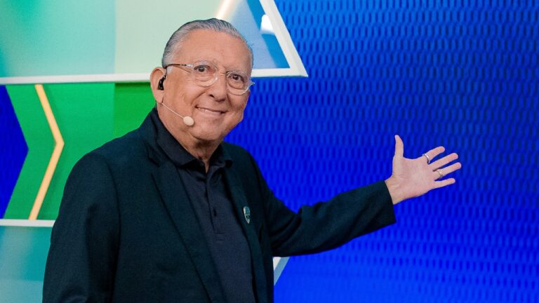 Galvão Bueno sorri e aponta para o cenário azul do programa Galvão F.C. usando microfone de cabeça no SBT