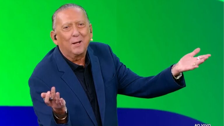 Foto do locutor esportivo Galvão Bueno, vestindo um terno azul, em um cenário moderno do SBT, com fundo verde e azul.