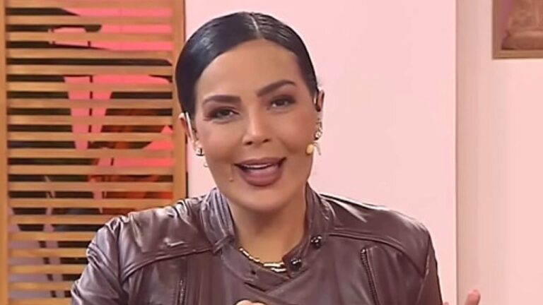Apresentadora com jaqueta marrom fala durante programa de TV em estúdio com iluminação rosa