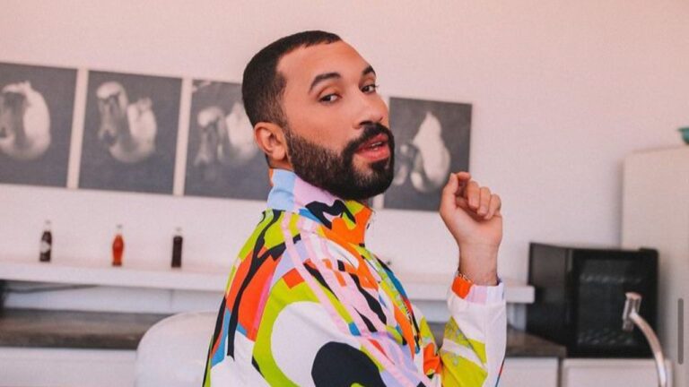 Gil do Vigor com barba e roupa colorida posa olhando para trás em ambiente interno com quadros na parede