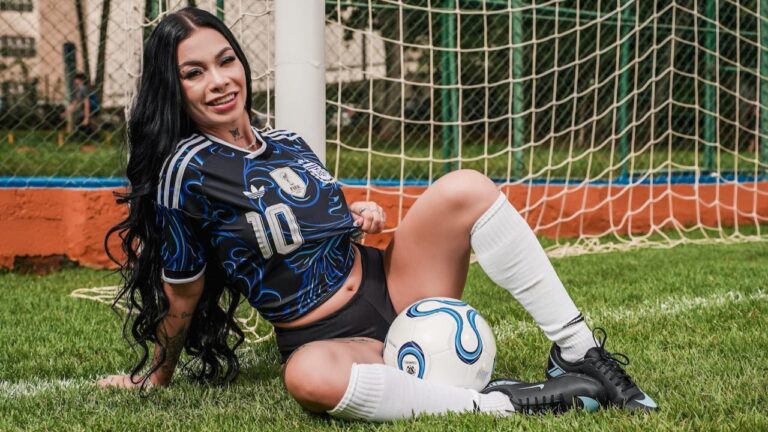 Gisa Custolli posa em campo de futebol com uniforme esportivo e bola diante da rede do gol