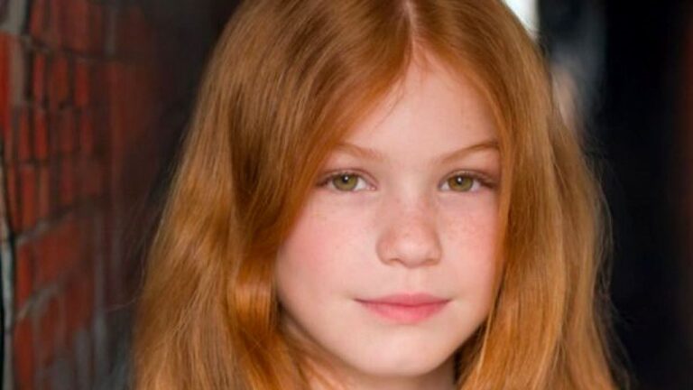Menina ruiva de olhos claros posa em retrato com expressão séria diante de fundo escuro