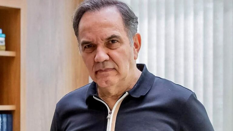 Homem de camisa polo preta olha sério para a câmera em ambiente interno com estante ao fundo