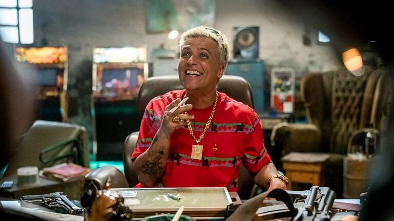 Homem de cabelo platinado e camisa vermelha sorri sentado em escritório com armas e objetos sobre a mesa