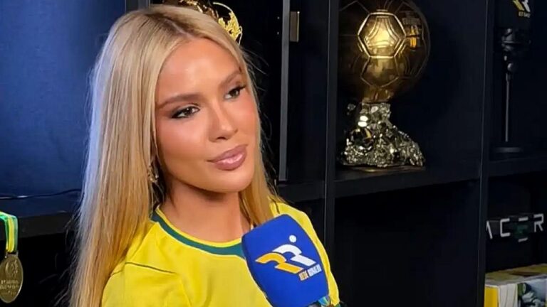 Mulher loira com camisa da seleção brasileira concede entrevista diante de prateleira com troféus
