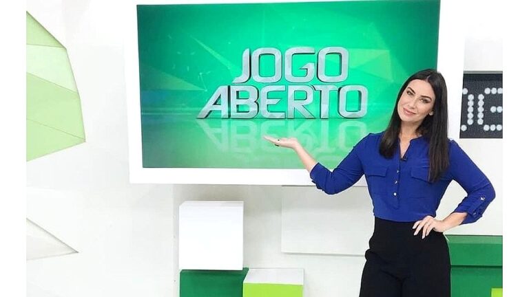 Larissa Erthal posa no estúdio do Jogo Aberto apontando para o logotipo do programa esportivo da Band