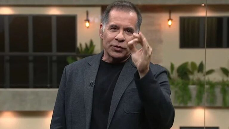 Apresentador de terno escuro gesticula durante fala em programa de TV com cenário moderno ao fundo