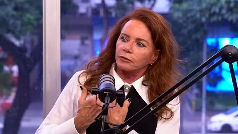 Mulher ruiva fala ao microfone durante entrevista em podcast com janela ao fundo