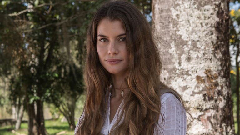 Alinne Moraes como Lívia encostada em árvore em cena da novela Além do Tempo ao ar livre