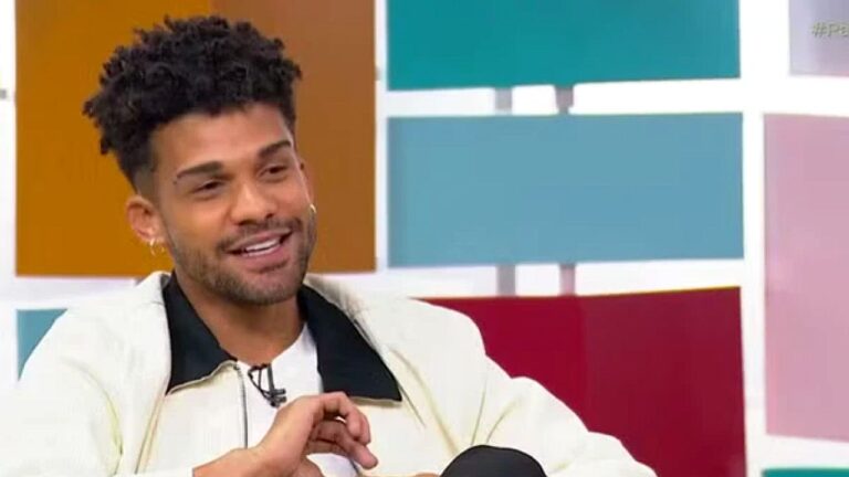 Homem sorri durante entrevista em programa de TV sentado em sofá no estúdio