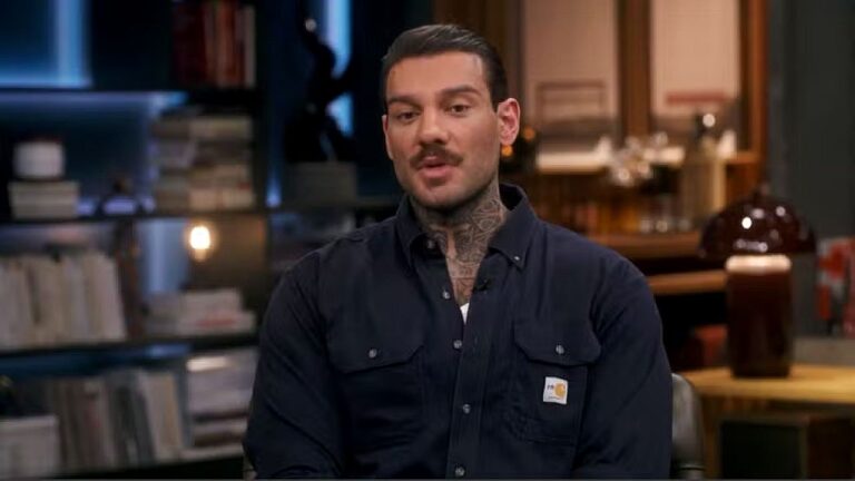 Homem com tatuagens no pescoço fala sentado em estúdio com estante de livros e iluminação aconchegante ao fundo